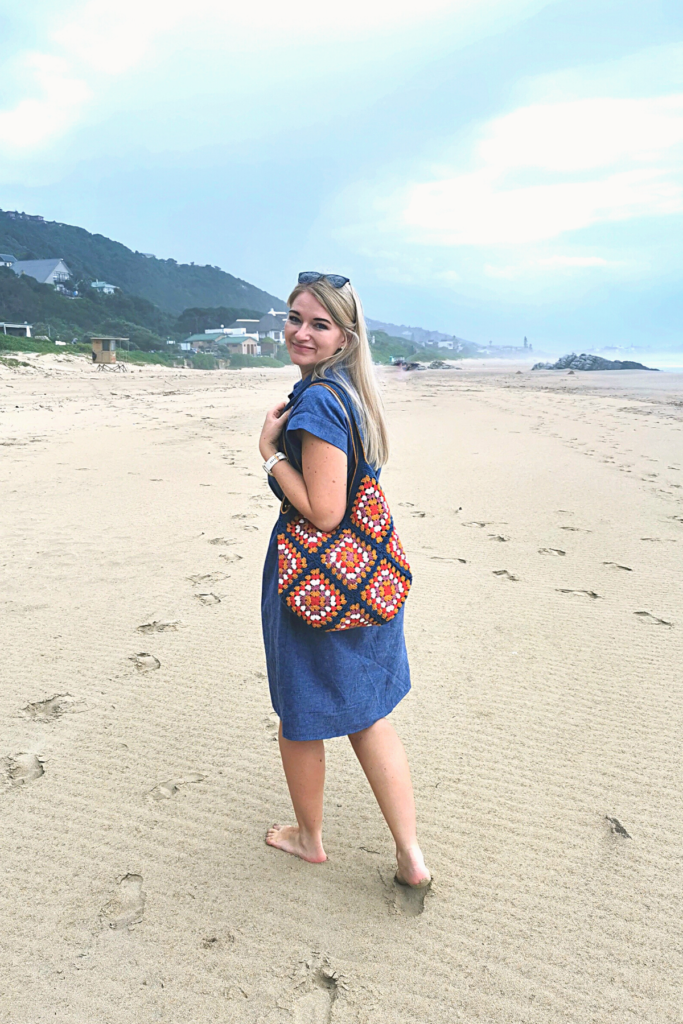 a bond woman standing on a beach holding a blue granny square tote bag.